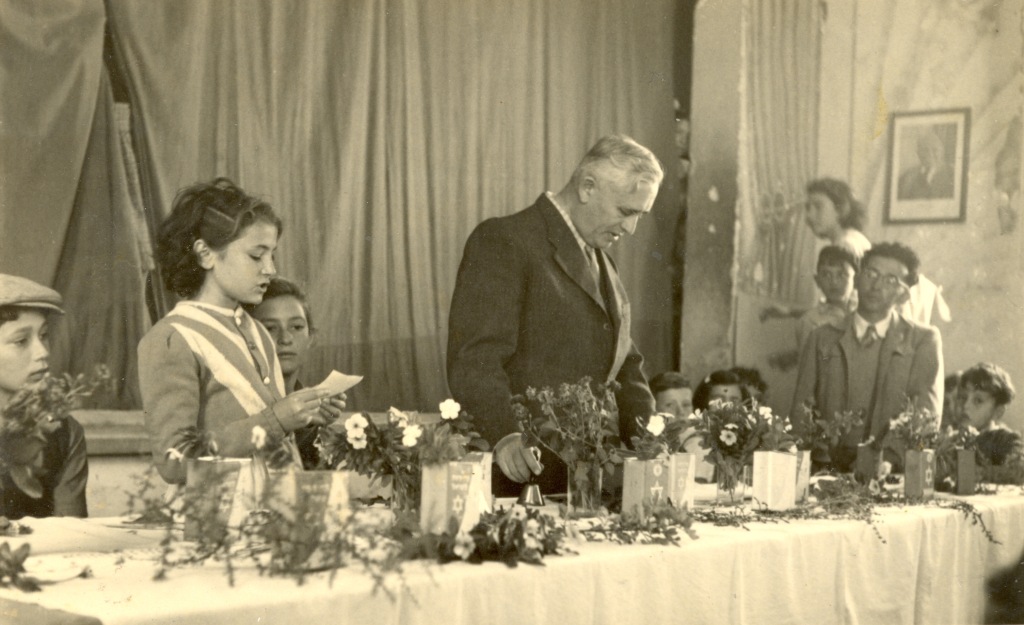 1947 טקס ראש החודש לכבוד הקופסה הכחולה של קק"ל באולם בית העם. במרכז עומד המנהל זלטין-זיו. התלמידה העומדת וקוראת היא אורה סולוביצ'יק.